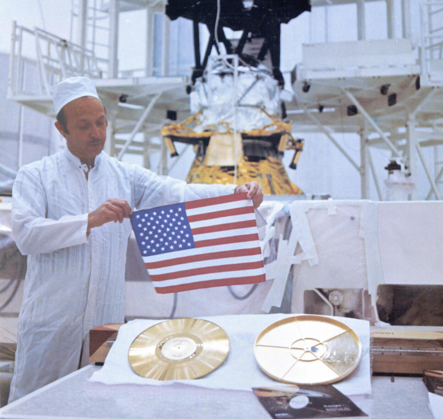 Archive image of a NASA employee holding up an American flag beside a golden 12" vinyl record in the 1970s.