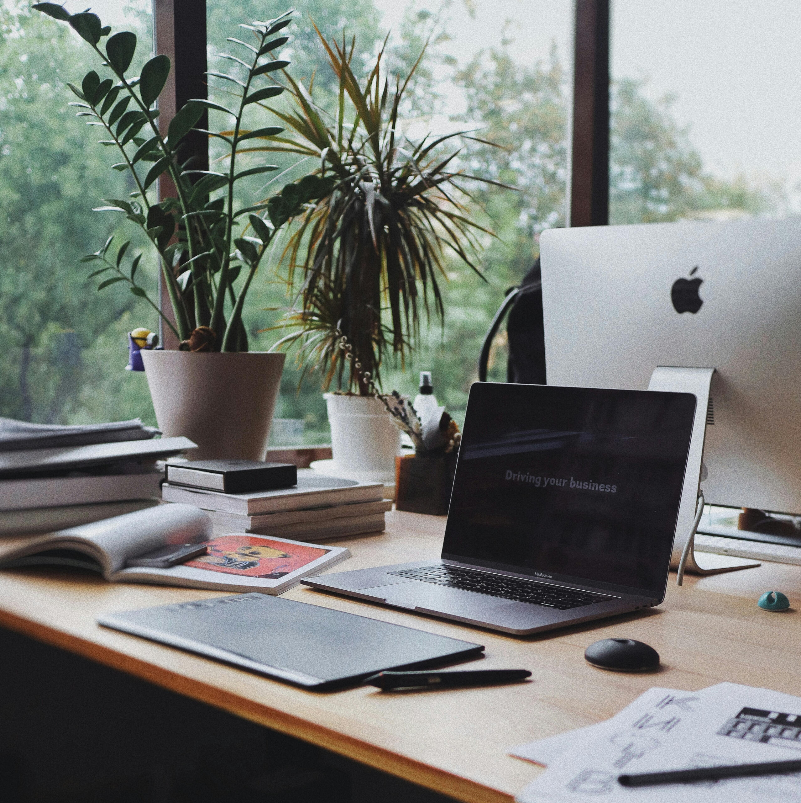 Photo of a laptop on a desk with notebooks and plants.